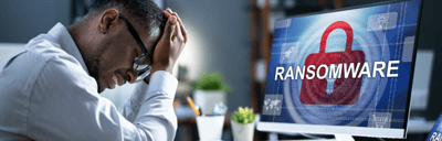 A man is holding his head in front of a computer screen with the word ransomware on it.